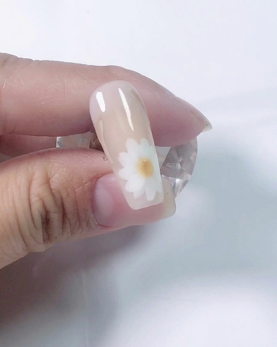 Un plano detalle de una manicurista usando un pincel fino para pintar pétalos de margarita blancos en una uña de práctica nude.