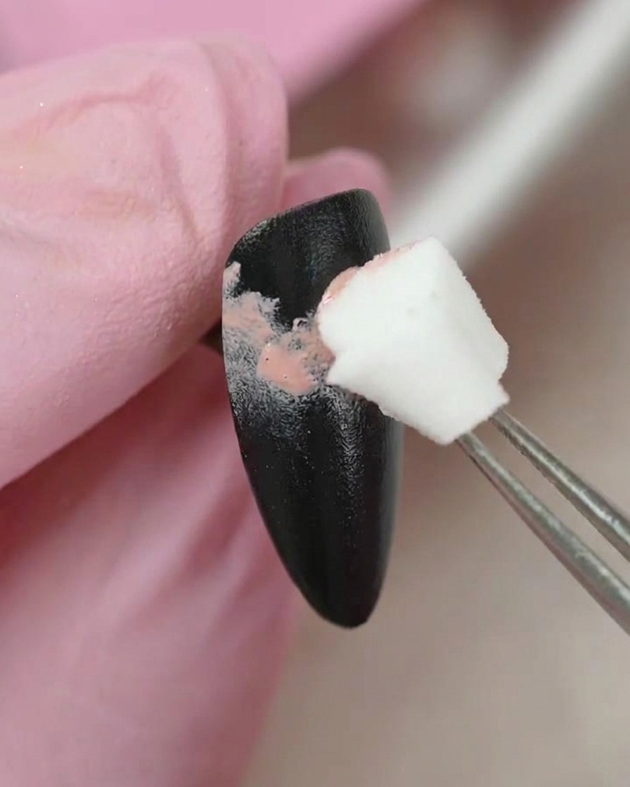 Un par de pinzas sosteniendo un pequeño aplicador de esponja, aplicando suavemente color de gel rosa sobre la superficie de una uña pintada de negro.
