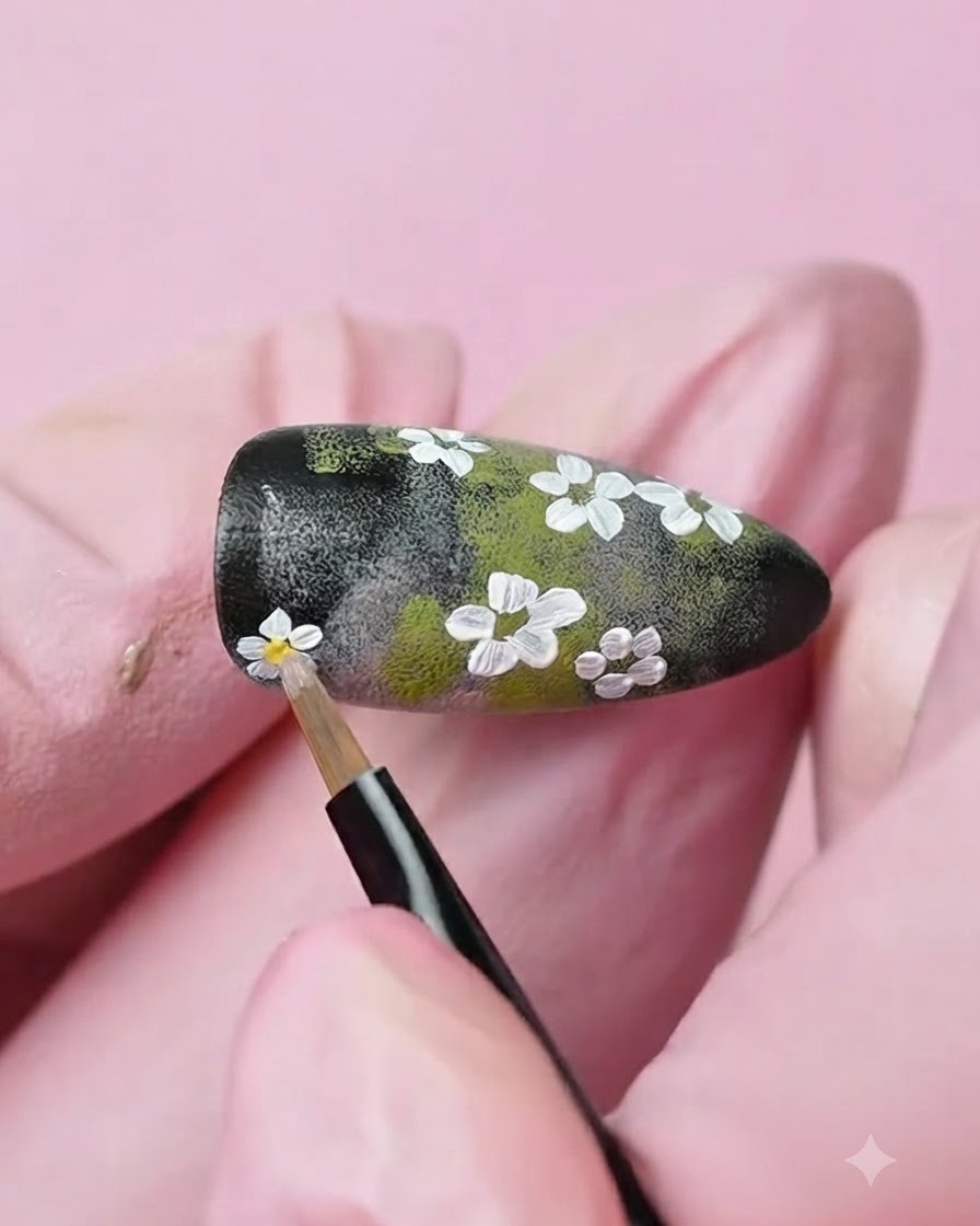 Un pincel de detalle fino aplicando un punto de gel amarillo en el centro de una flor blanca pintada en la uña.