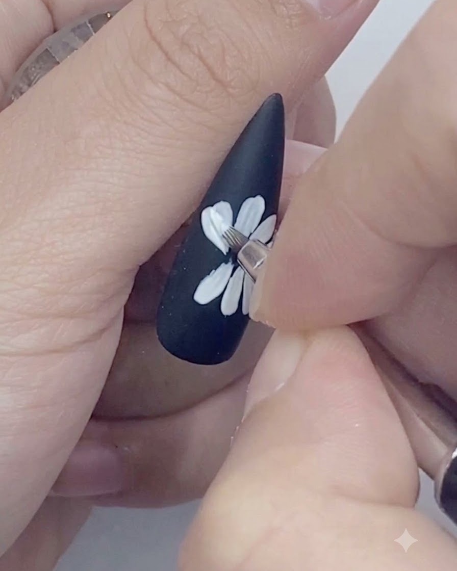 Un pincel delineador fino trazando los bordes exteriores de los pétalos de margarita blancos pintados sobre una uña negra.