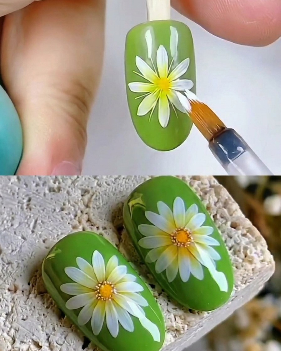 Un primer plano de un pincel aplicando una capa superior transparente brillante sobre un diseño de arte de uñas de margarita blanca en una uña verde.