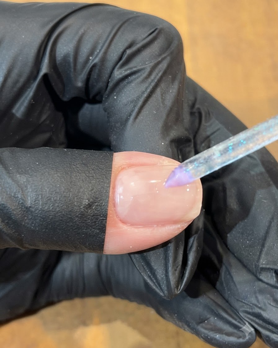 Una mano con guante usando una herramienta de pincel fino para colocar un punto de gel azul junto a una sección morada en una uña.