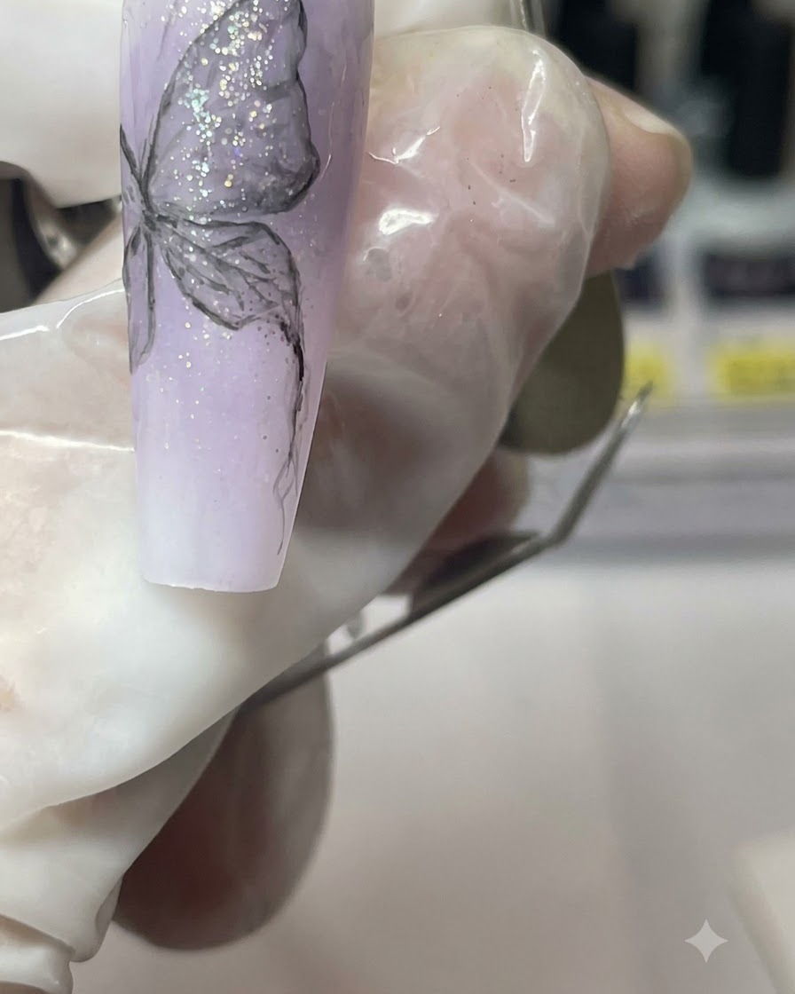 Un primer plano de un dedo con manicura sosteniendo la punta de uña morada mientras detalla el cuerpo de la mariposa.