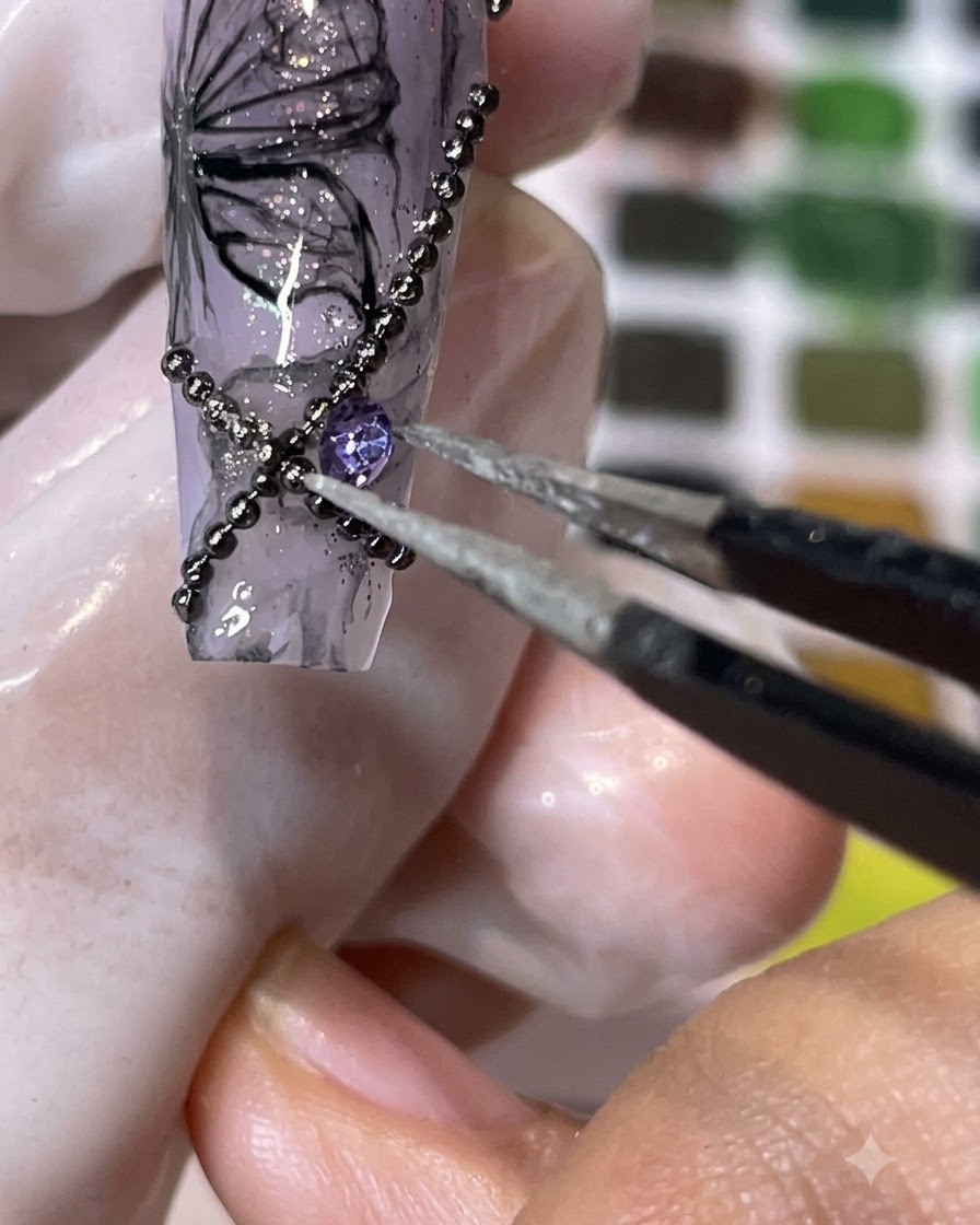Un par de pinzas colocando un strass morado de base cónica sobre una superficie de uña decorada.