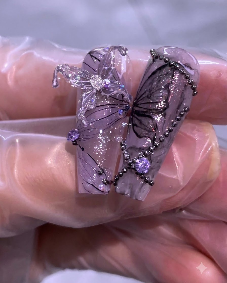 Primer plano de arte de uñas completado en tonos morado y negro con detalles de mariposas, pedrería y delicadas líneas de hilos.