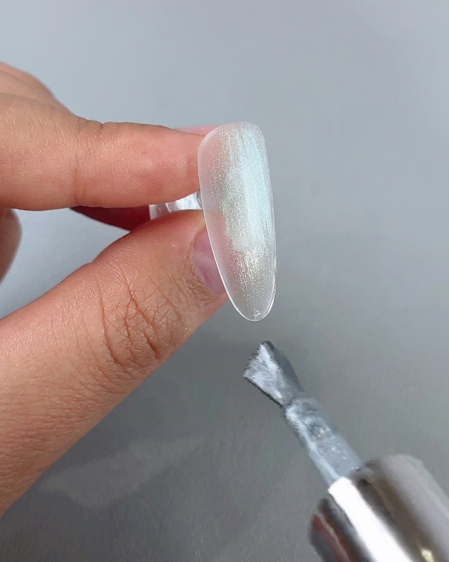 Un pincel de uñas aplicando una capa de gel cat eye plateado brillante sobre la punta de una uña transparente.