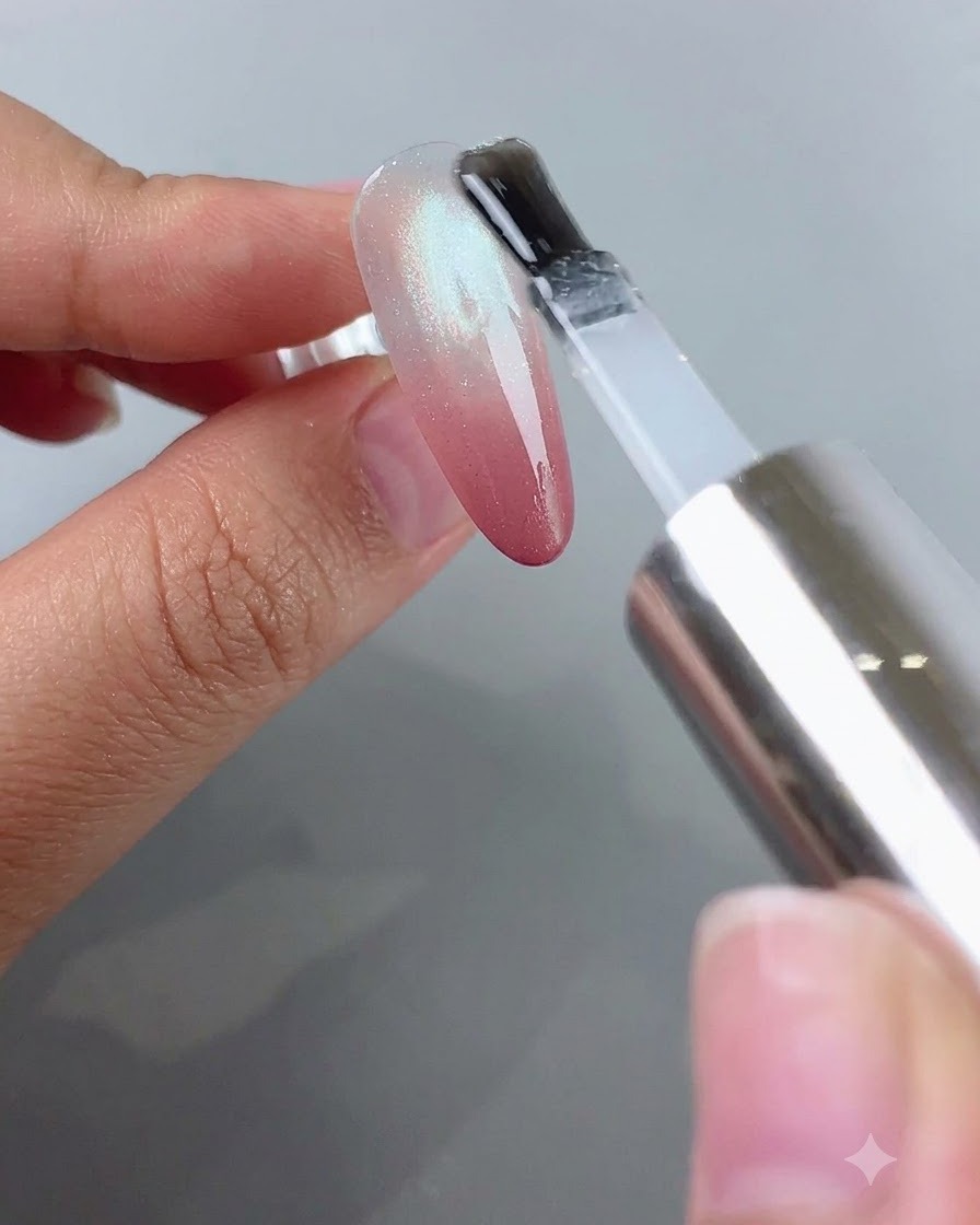 Un pincel aplicando top coat transparente sobre una uña que tiene un efecto blush degradado rosa y cat eye plateado.