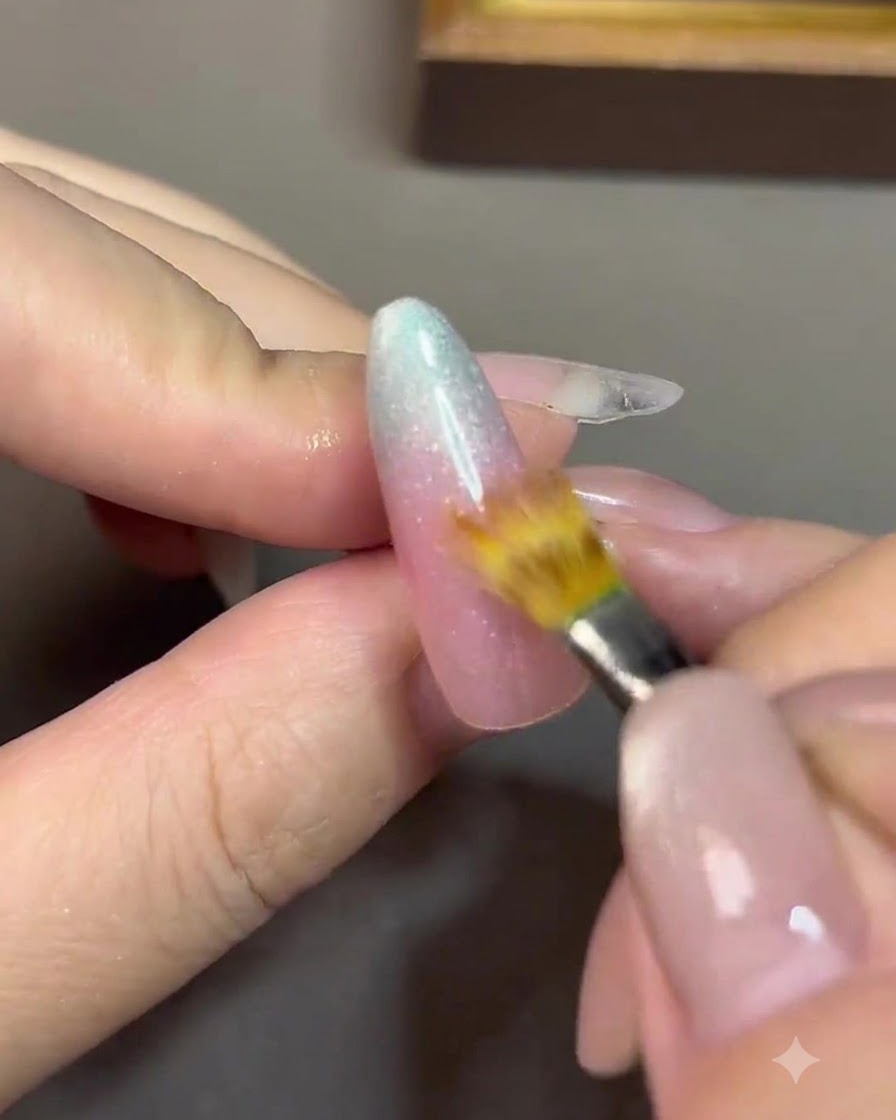 Un pincel de detalle fino difuminando la línea de transición entre un gel rosa nude y un gel azul en una uña.