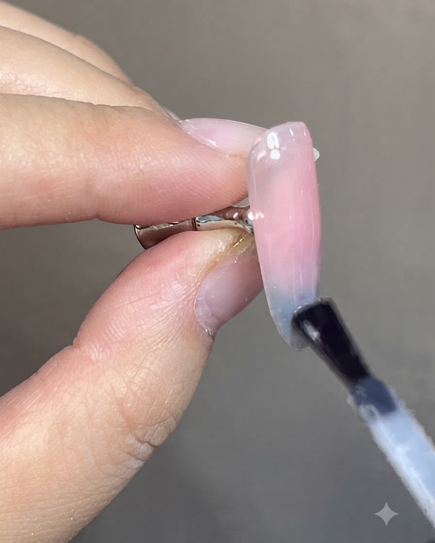 Un pincel aplicando una capa superior brillante transparente sobre un diseño de uñas degradado cat-eye azul y rosa terminado.