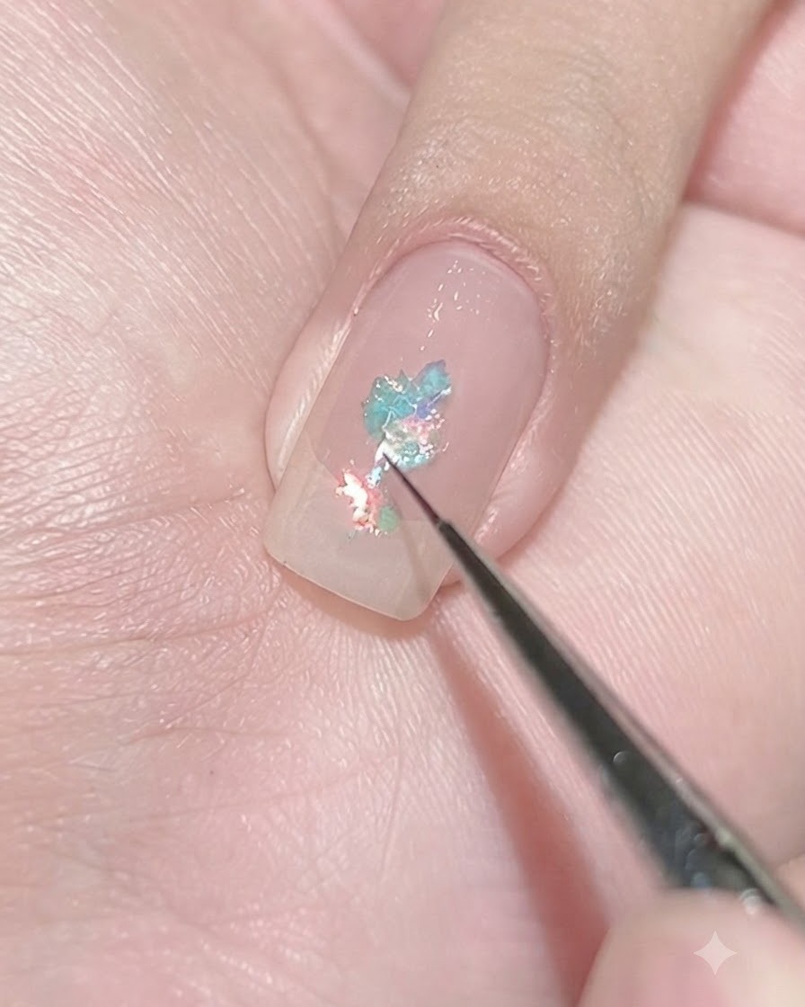 Un pincel delineador fino colocando pequeñas escamas celestes sobre una uña con base transparente para crear una agrupación concentrada.