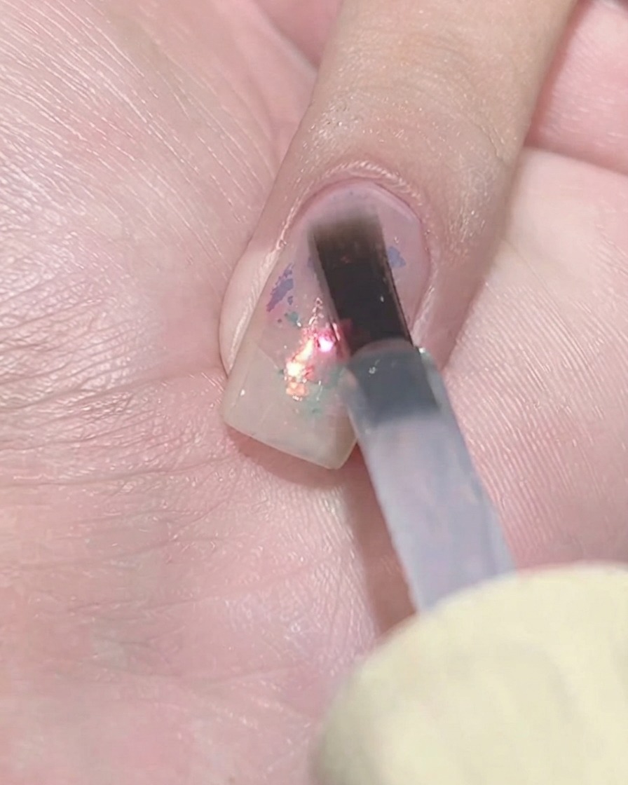 Un pincel plano para gel aplicando a toquecitos una capa de esmalte morado translúcido sobre un grupo de escamas texturizadas en una uña.