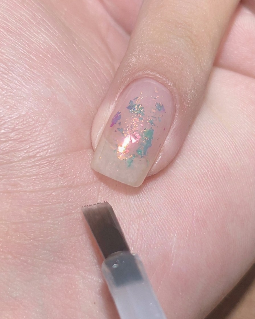 Un pincel plano para gel suspendido cerca de una uña transparente con escamas encapsuladas, listo para aplicar esmalte morado translúcido.