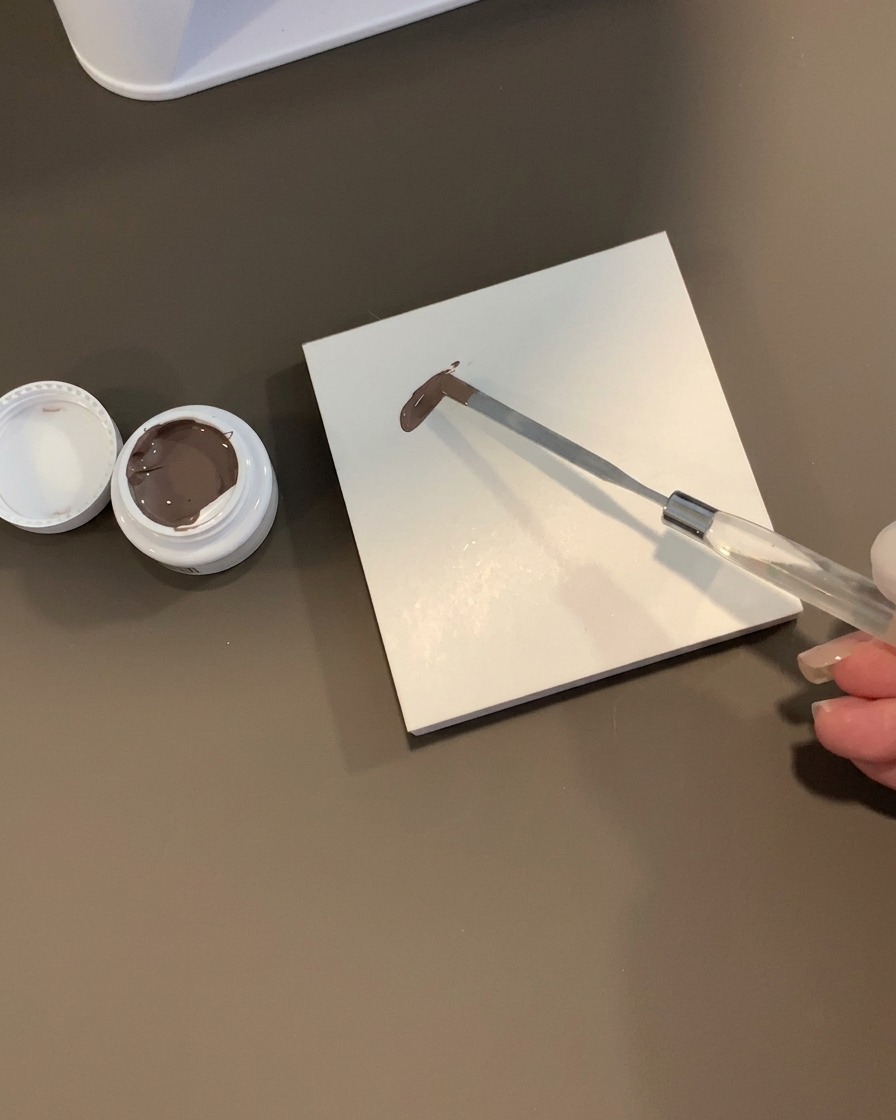Una espátula metálica con una gota de gel gris pardusco sobre una paleta cuadrada blanca junto a un tarro abierto.