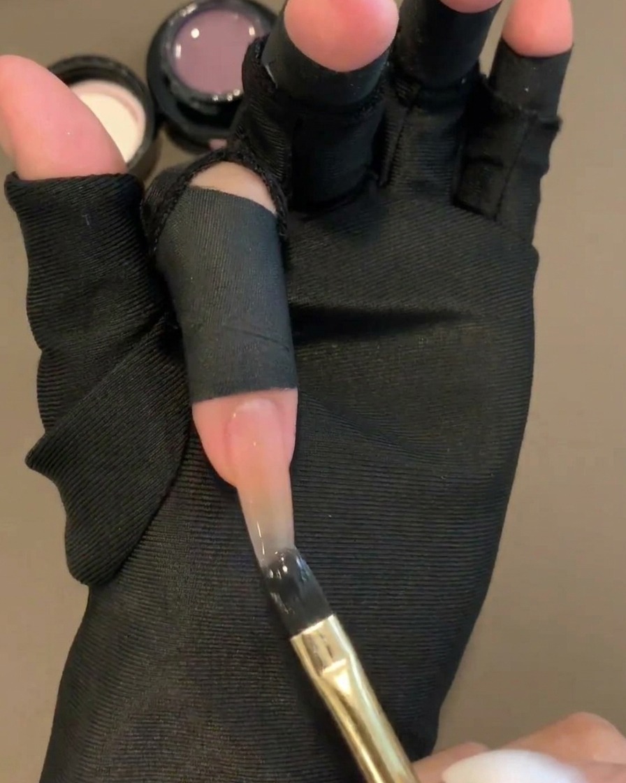 Un pincel de gel plano aplicando un color gel púrpura translúcido en la mitad superior de una uña con base nude.