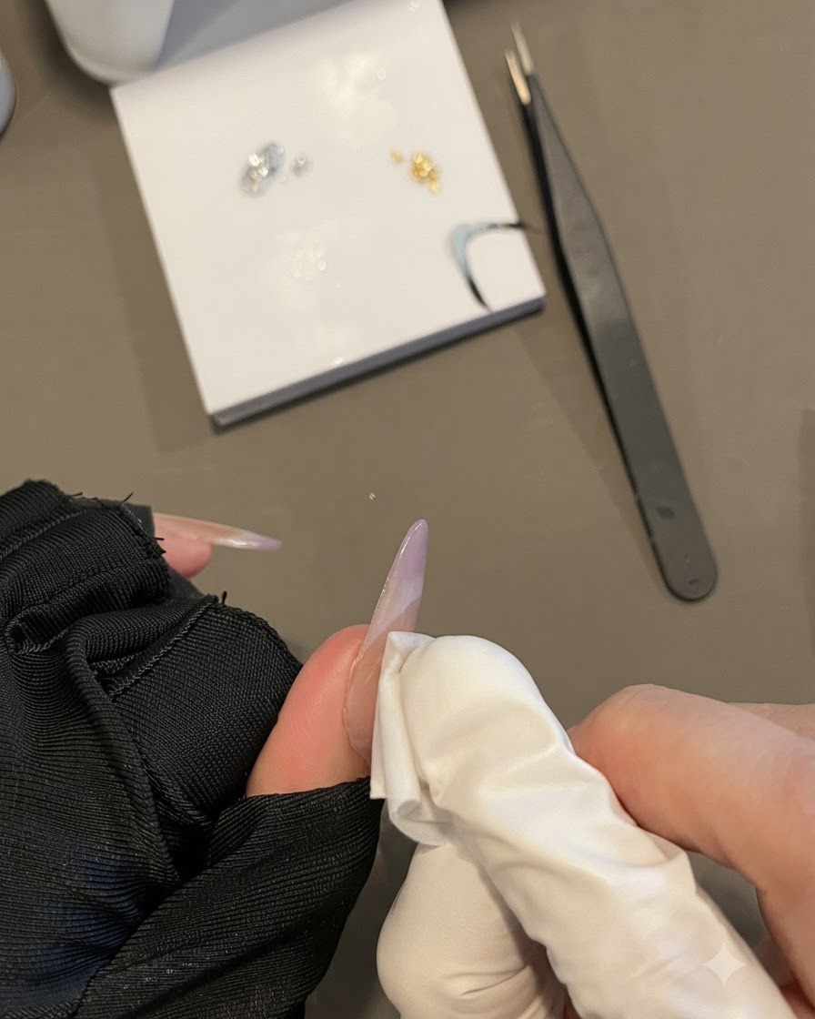 Una mano con guante limpiando la superficie de una uña con una toallita sin pelusa mientras sostiene una uña ovalada larga.