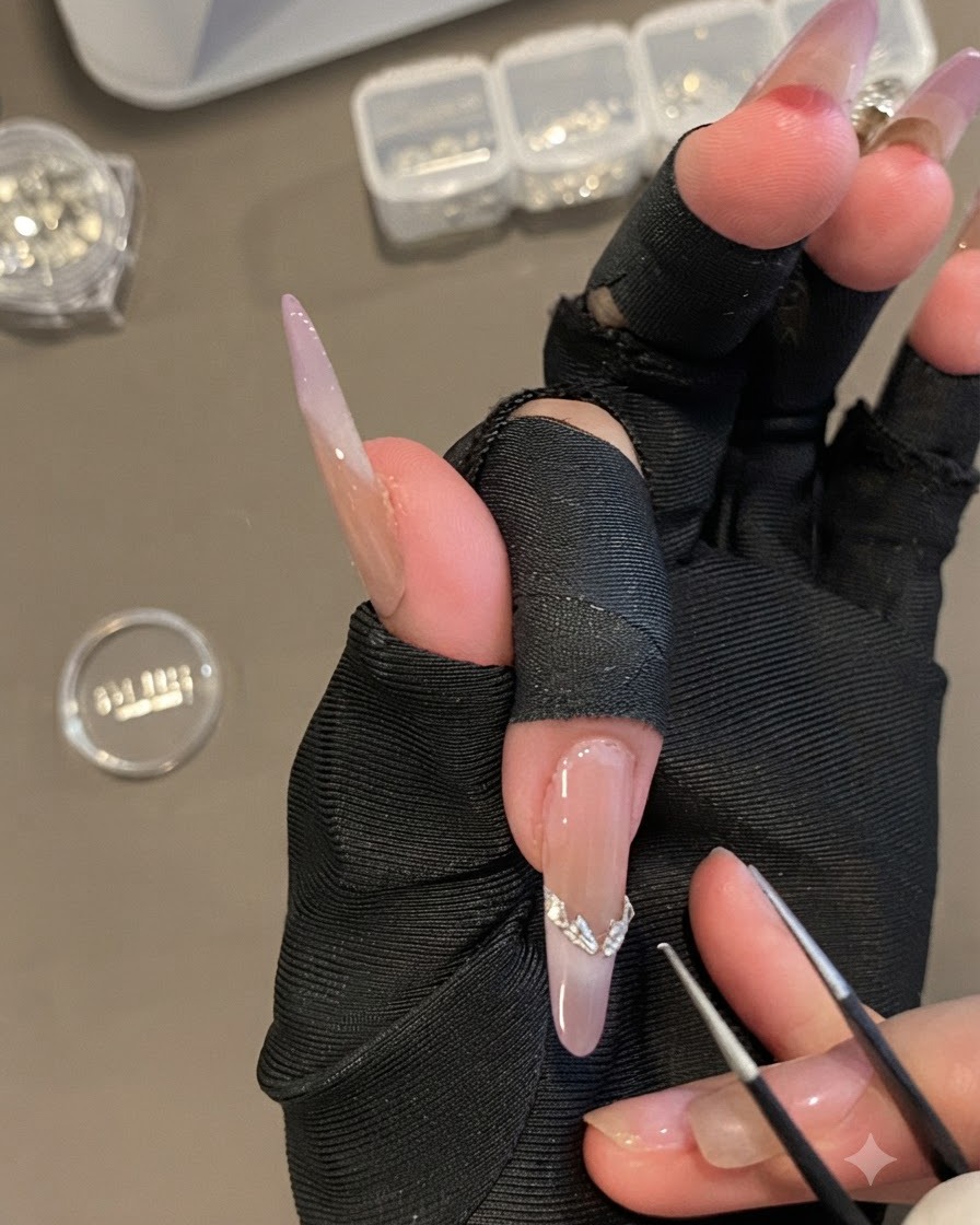 Una mano sosteniendo pinzas de precisión colocando una pegatina curva plateada de punta francesa en una uña ovalada larga.