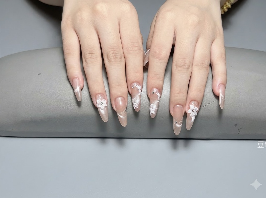 Un primer plano de uñas largas almendradas con flores blancas esculpidas en 3D y pequeños cristales plateados sobre una base nude brillante.
