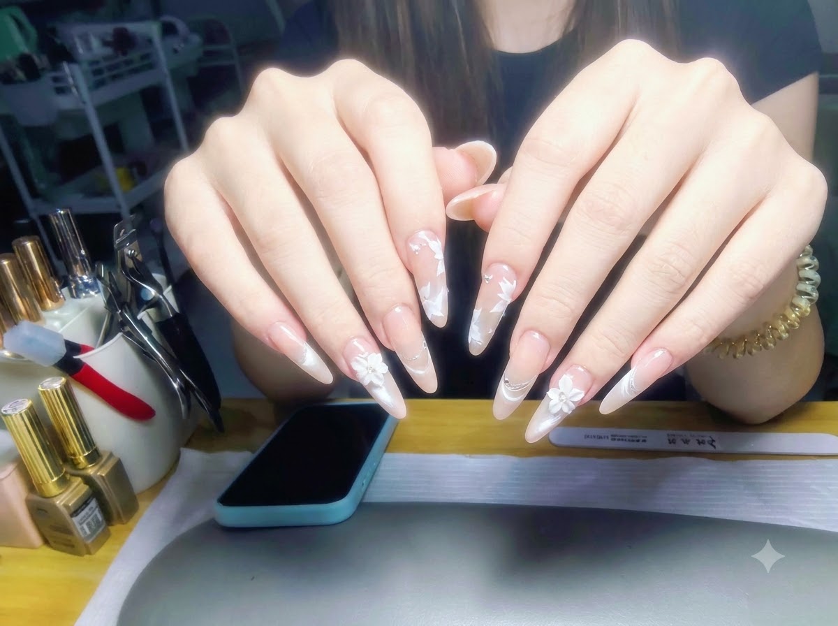 Ambas manos levantadas frente al fondo de una estación de trabajo, mostrando un juego completo de uñas brillantes con degradado nude perlado y arte floral blanco.