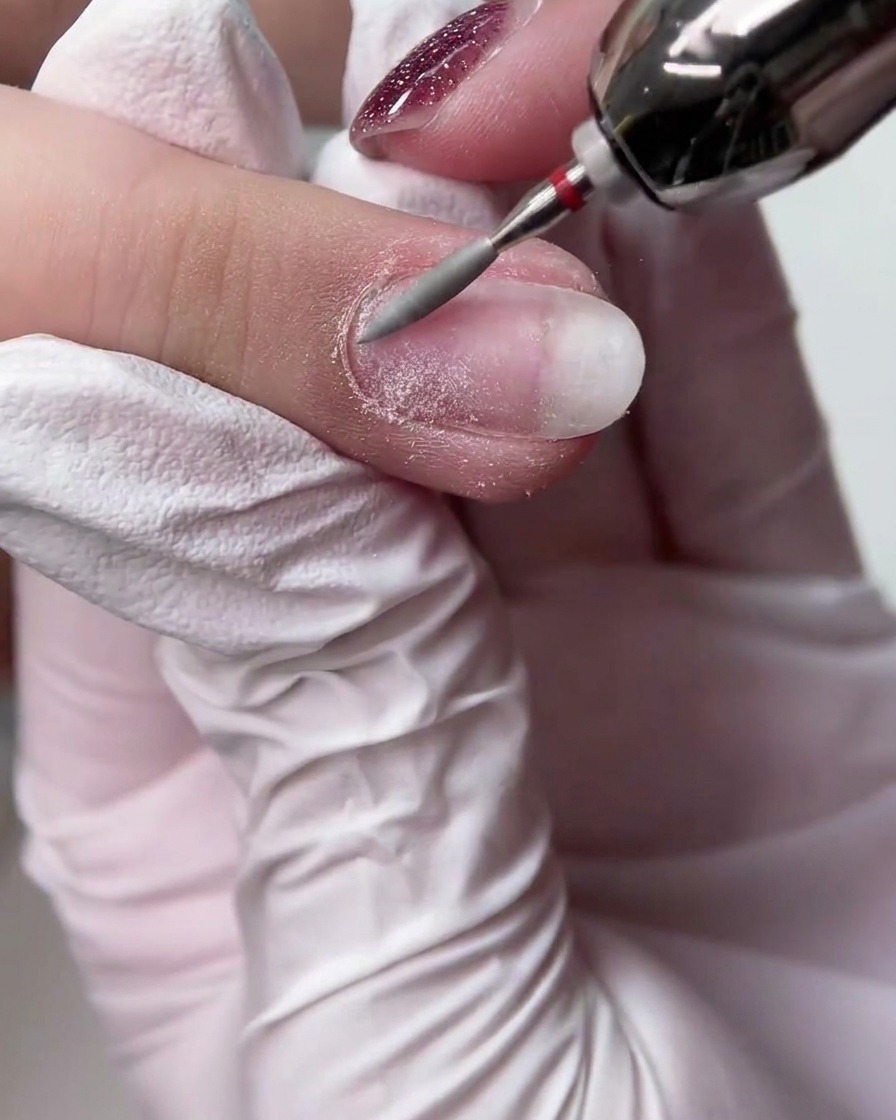 Una mano con guante usando un torno eléctrico con una fresa cónica larga para alisar la superficie de una uña natural.