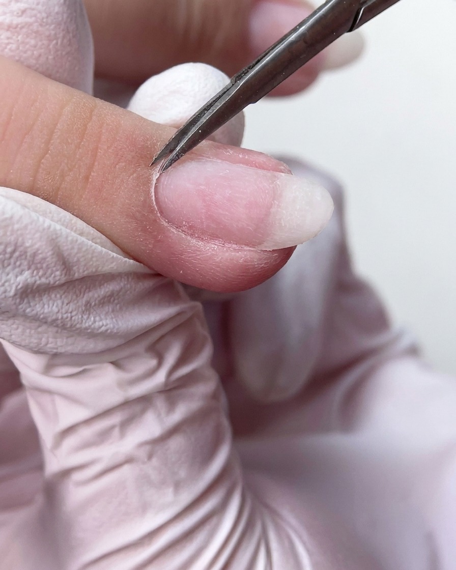 Tijeras de manicura finas recortando ordenadamente el exceso de tejido de cutícula muerta en la base de la uña.