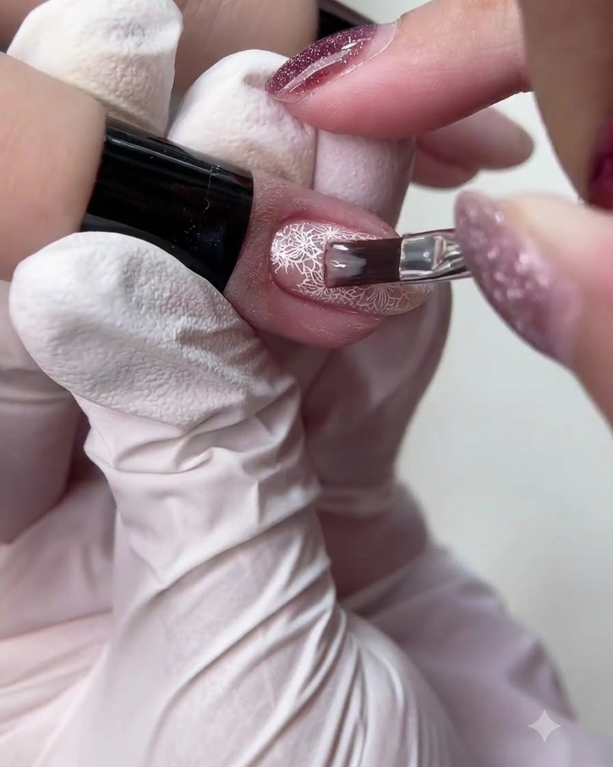 Un pincel de uñas pequeño aplicando un esmalte de gel nude translúcido sobre un diseño detallado de arte de uñas de encaje blanco.