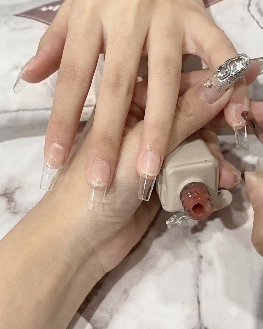 Un pincel de uñas aplicando una capa fina de gel nude translúcido sobre una uña natural larga en forma de ataúd.