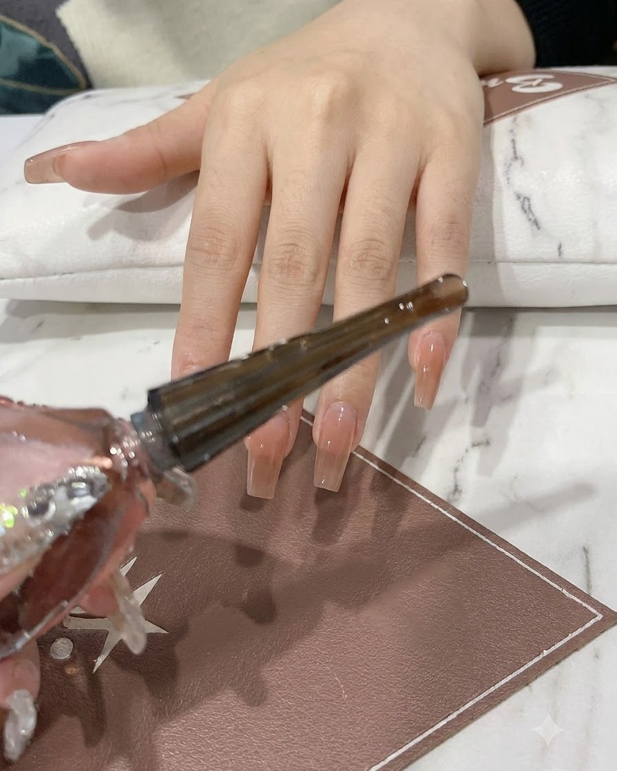 Una mano sosteniendo un frasco de gel rosa y plateado brillante con el aplicador de pincel extendido.