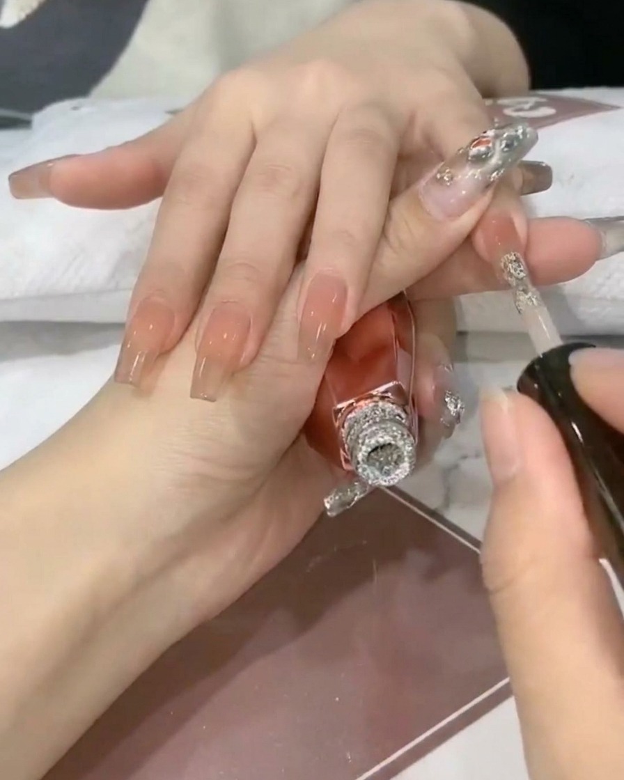 Un pincel de uñas depositando gel con purpurina plateada brillante en la punta de una uña con base nude translúcida.