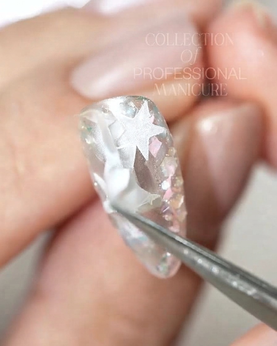 Un par de pinzas despegando piezas intrincadas de pegatinas de estrellas blancas de una uña brillante.