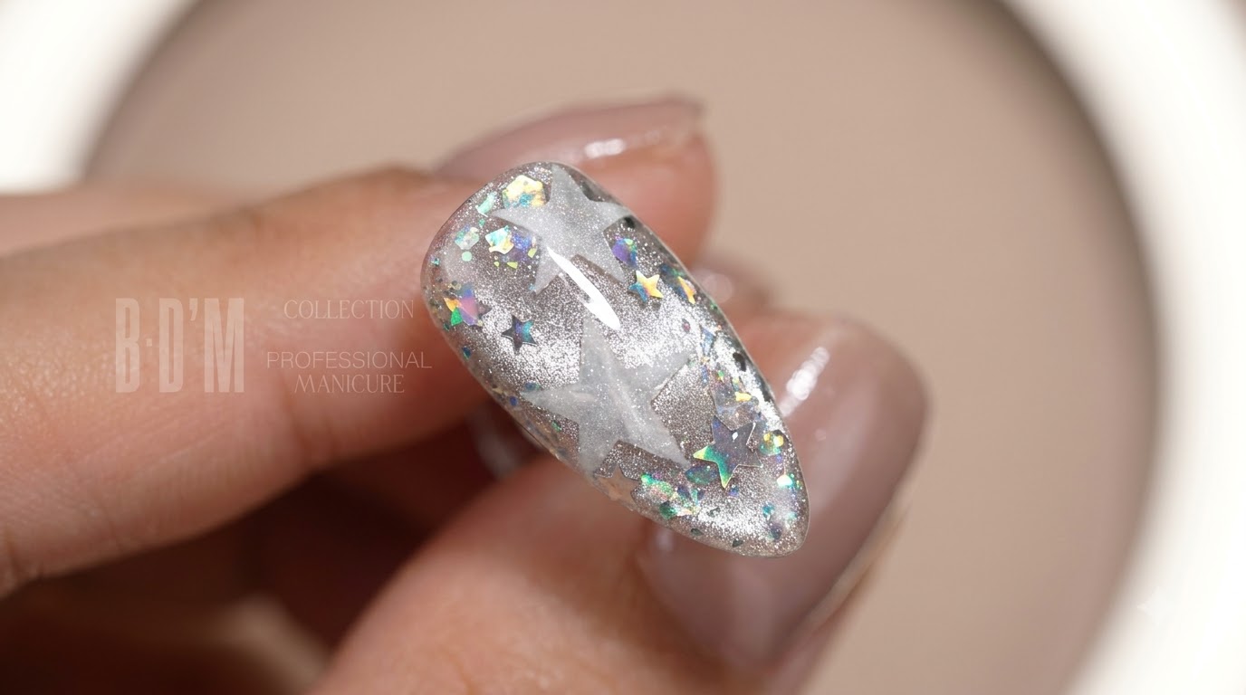 Primer plano de una uña almendrada brillante terminada con efectos de ojo de gato plateado, lentejuelas de estrellas plateadas y holográficas transparentes, y motivos de estrellas blancas en capas.