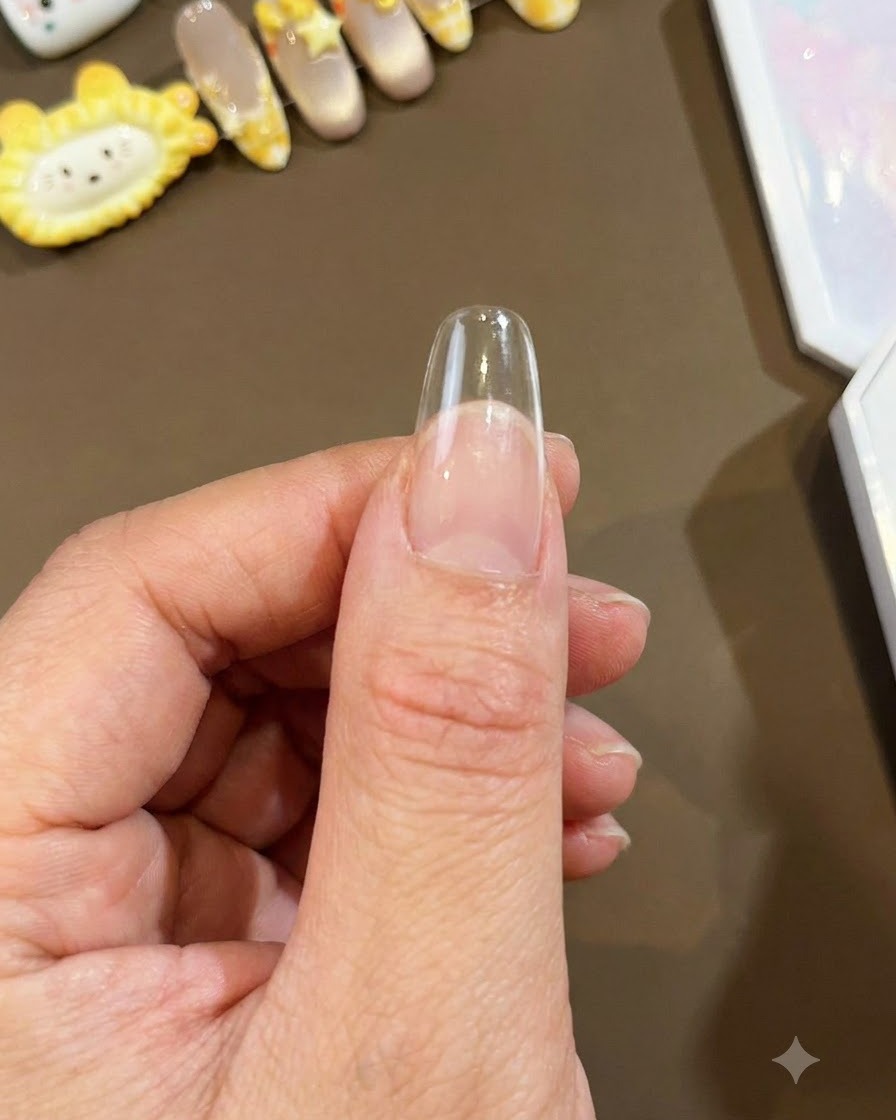 Un primer plano de un tip de uña largo y transparente sostenido contra un pulgar, listo para la aplicación de arte en uñas.