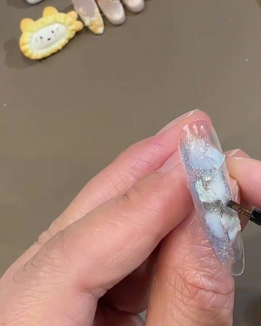 Un pincel fino de detalle aplicando polvo de hadas brillante blanco sobre una uña postiza transparente decorada con piezas de concha azul.