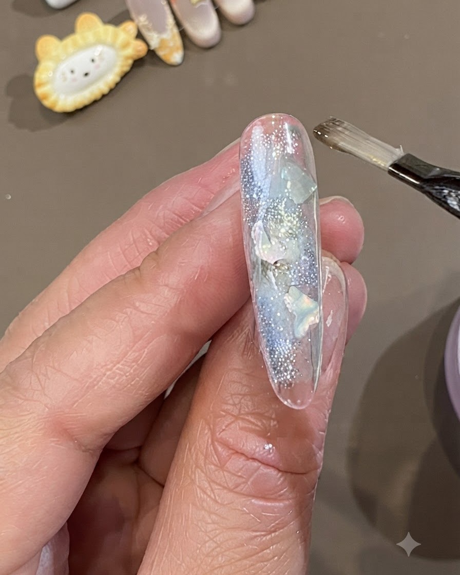 Un pincel de gel plano aplicando una capa gruesa de gel de encapsulado suave transparente sobre una uña decorada con piezas de concha y purpurina.