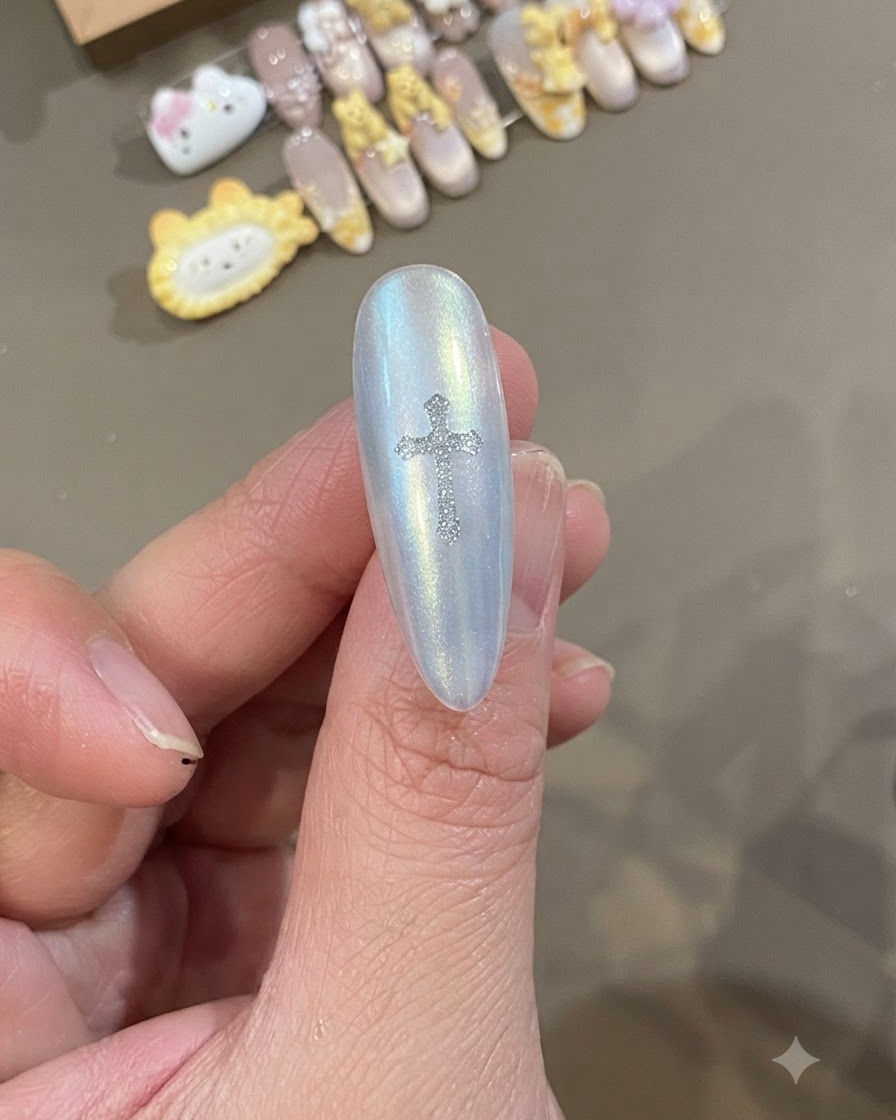 Un primer plano de una uña blanca perlada con una pegatina brillante de cruz de Corazón Cromado plateado colocada directamente en el centro.