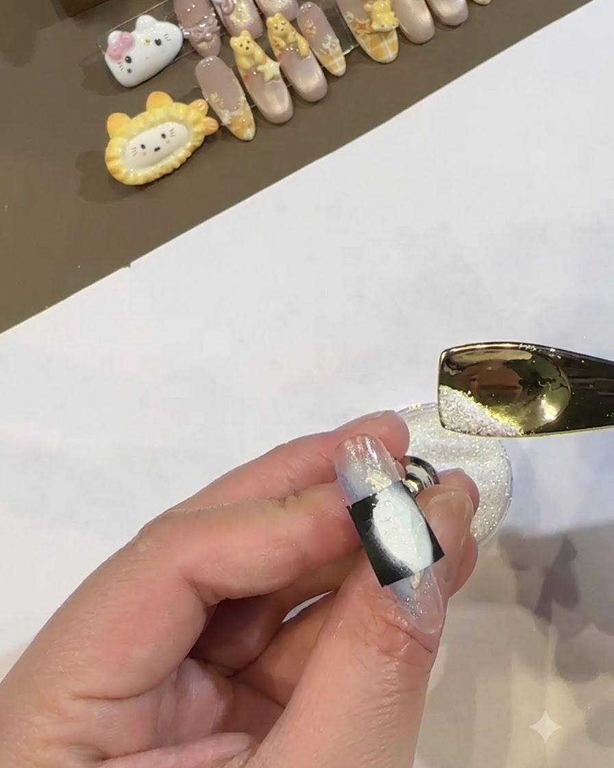 Una cuchara de metal dorada vertiendo polvo blanco brillante sobre el área de gel blanco húmedo enmarcada por una plantilla negra en una uña.