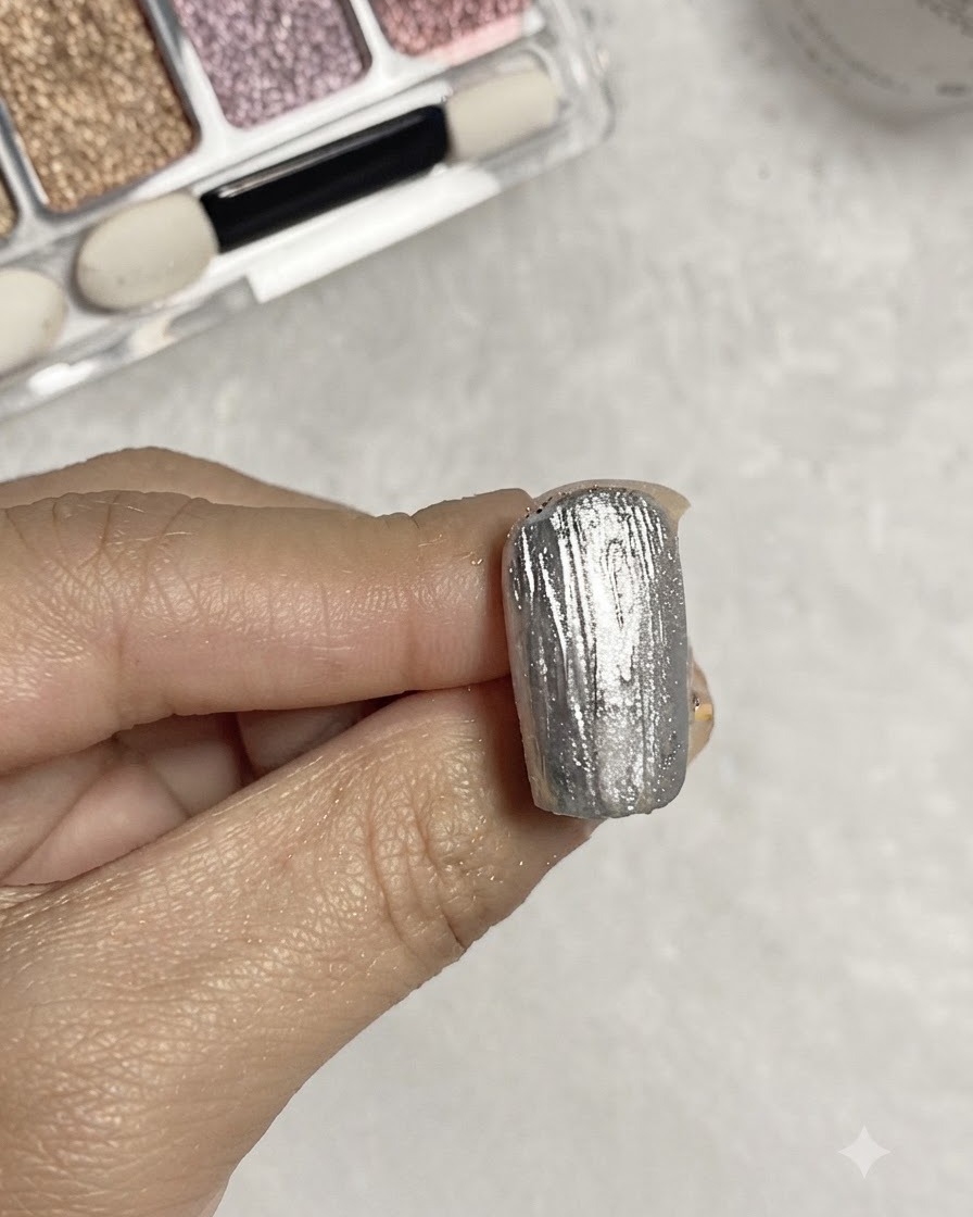 Un pincel aplicando gel base transparente sobre una superficie de uña con textura metálica plateada.