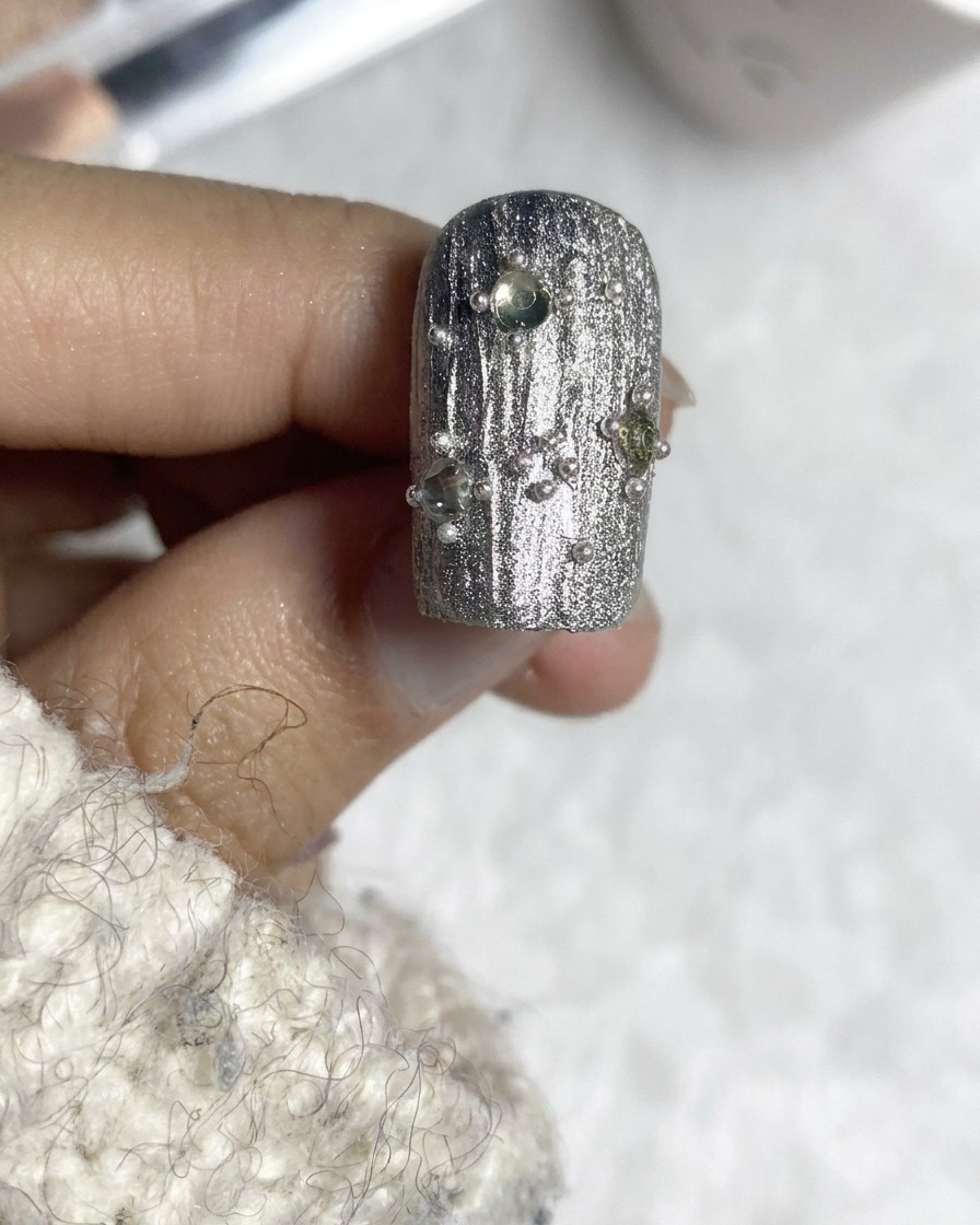 Un pincel de capa superior finalizando un diseño de uñas texturizado plateado y adornado.