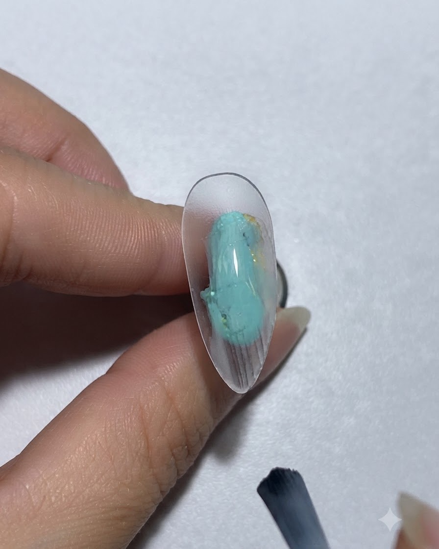 Un pincel aplicando gel de pintura en color azul verdoso sobre una uña.
