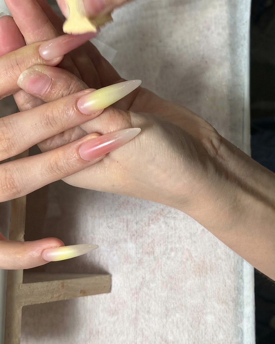 Una mano recibiendo trabajo con una esponja que mezcla tonos verde y rojo coral en uñas largas almendradas.