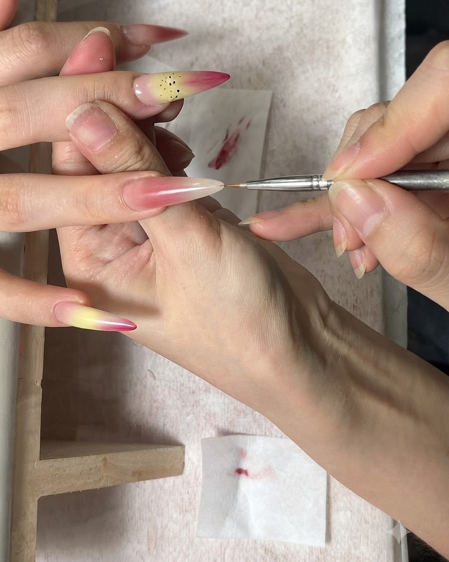 Un pincel fino de arte en uñas aplicando puntos marrones como estambres sobre una uña decorada con degradado.