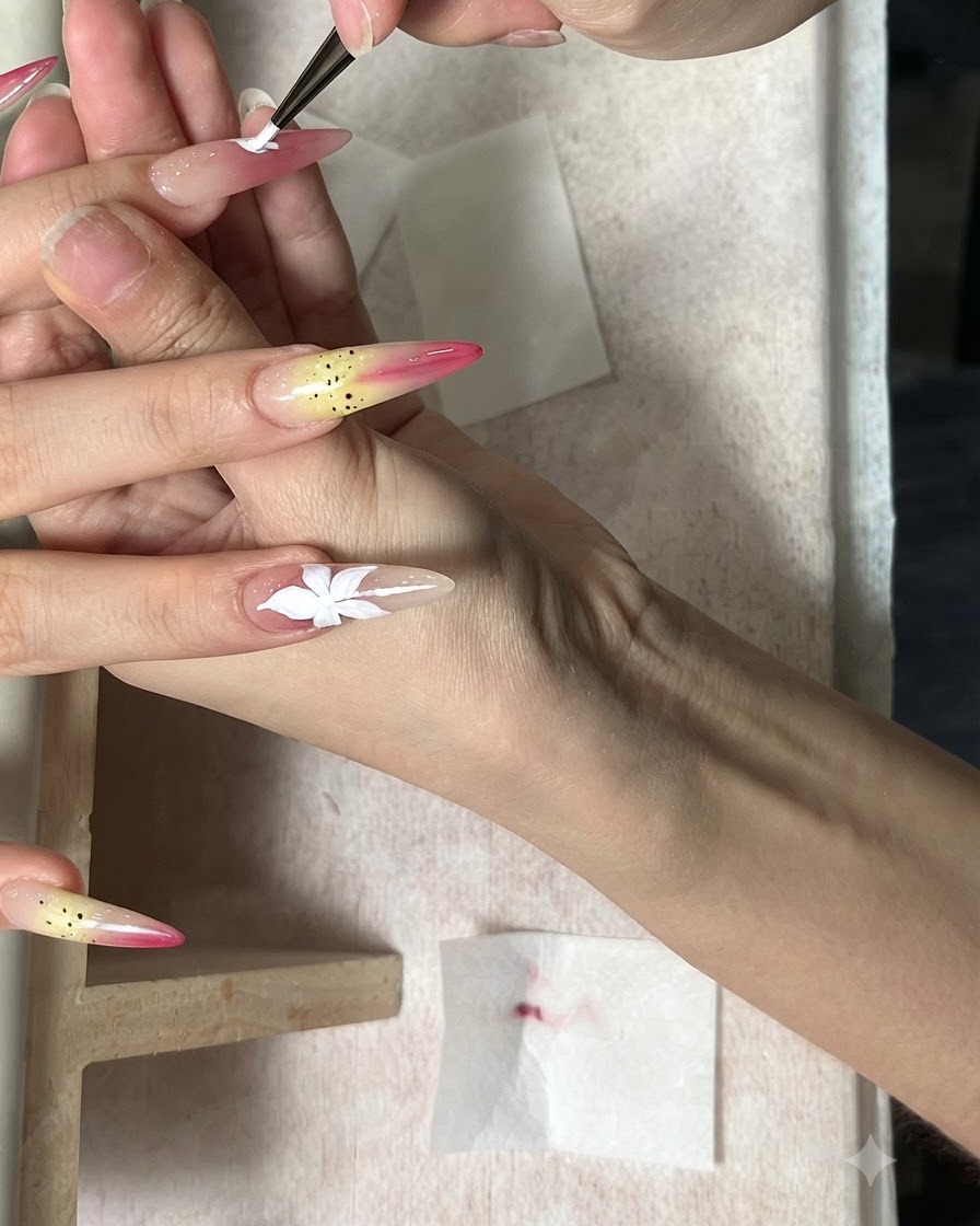 Un pincel fino pintando una flor blanca de cinco pétalos en una uña alargada color nude.