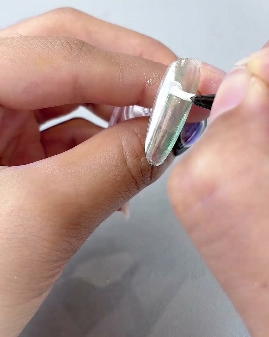 Un pincel fino aplicando gel de pintura blanco en pétalos curvos sobre una superficie plateada cromada.
