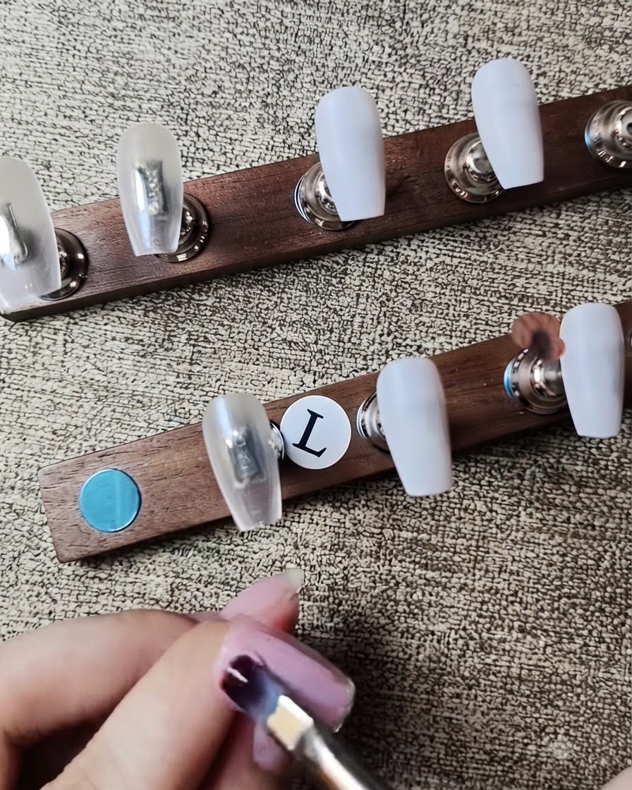 Un pincel plano aplicando esmalte en gel rosa nude translúcido sobre una uña postiza.
