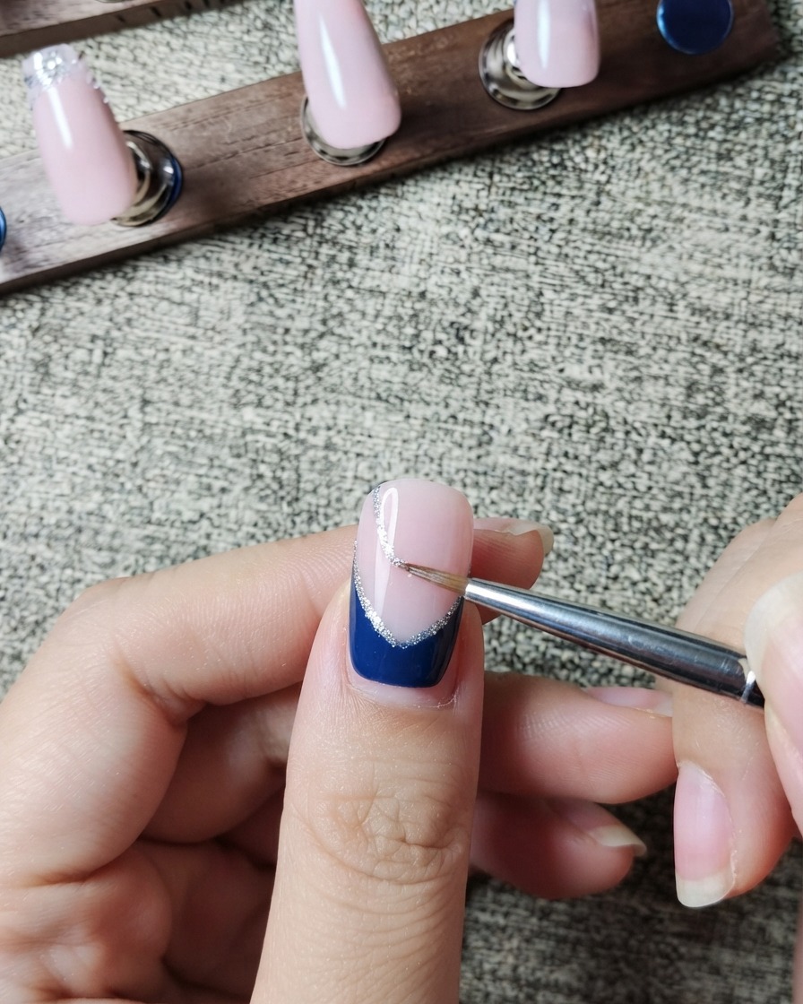 Un pincel delineador pintando cuidadosamente una fina línea de purpurina plateada a lo largo del borde interior de una punta francesa azul oscuro.