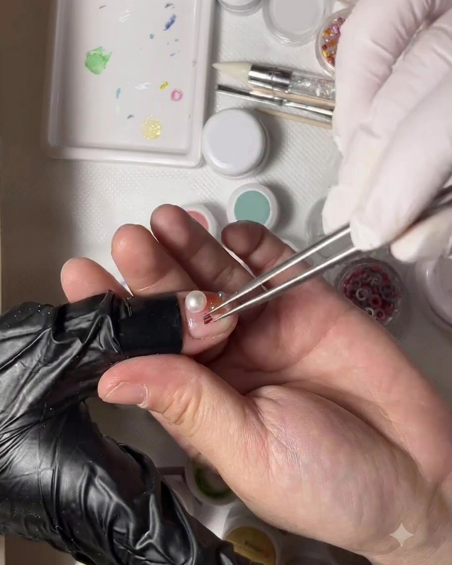 Una mano enguantada sosteniendo pinzas que colocan una pequeña cuenta dorada al lado de una perla en la uña decorada de un pulgar.