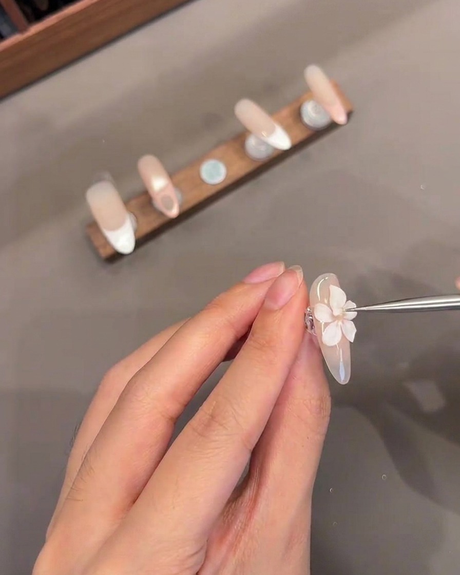 Un primer plano de una herramienta delgada añadiendo un pequeño centro de gel blanco a una flor 3D esculpida en una uña.