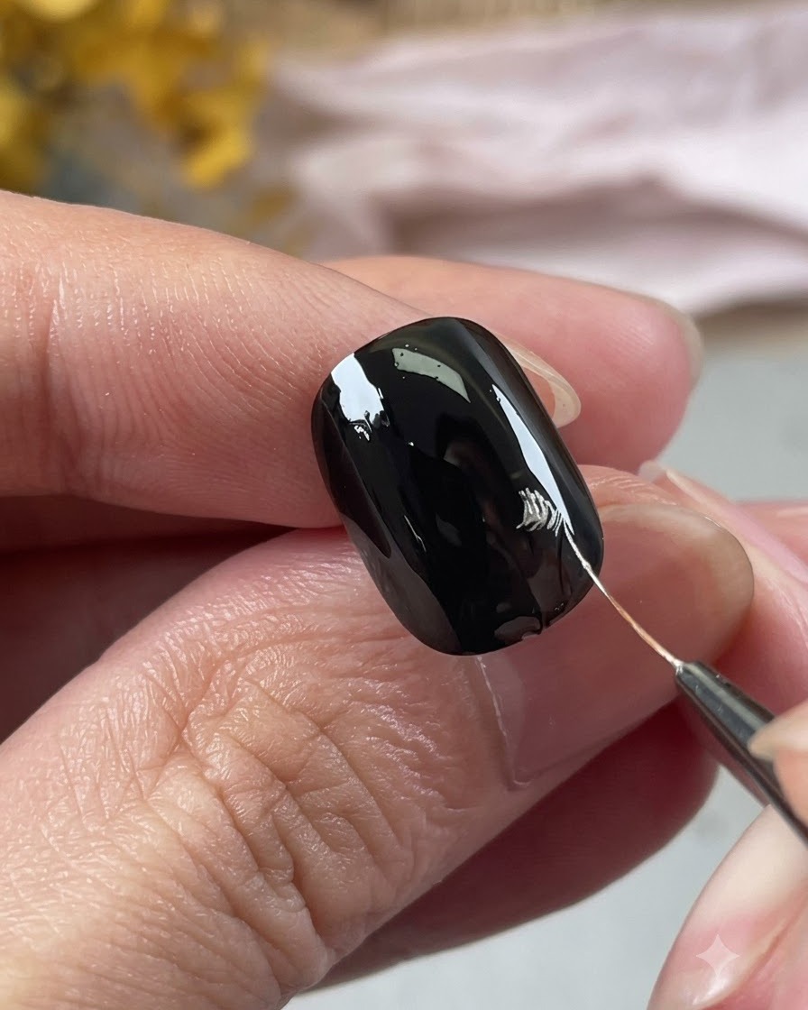 Primer plano de una mano usando un pincel fino para dibujar el contorno plateado de unos pétalos en una uña negra.