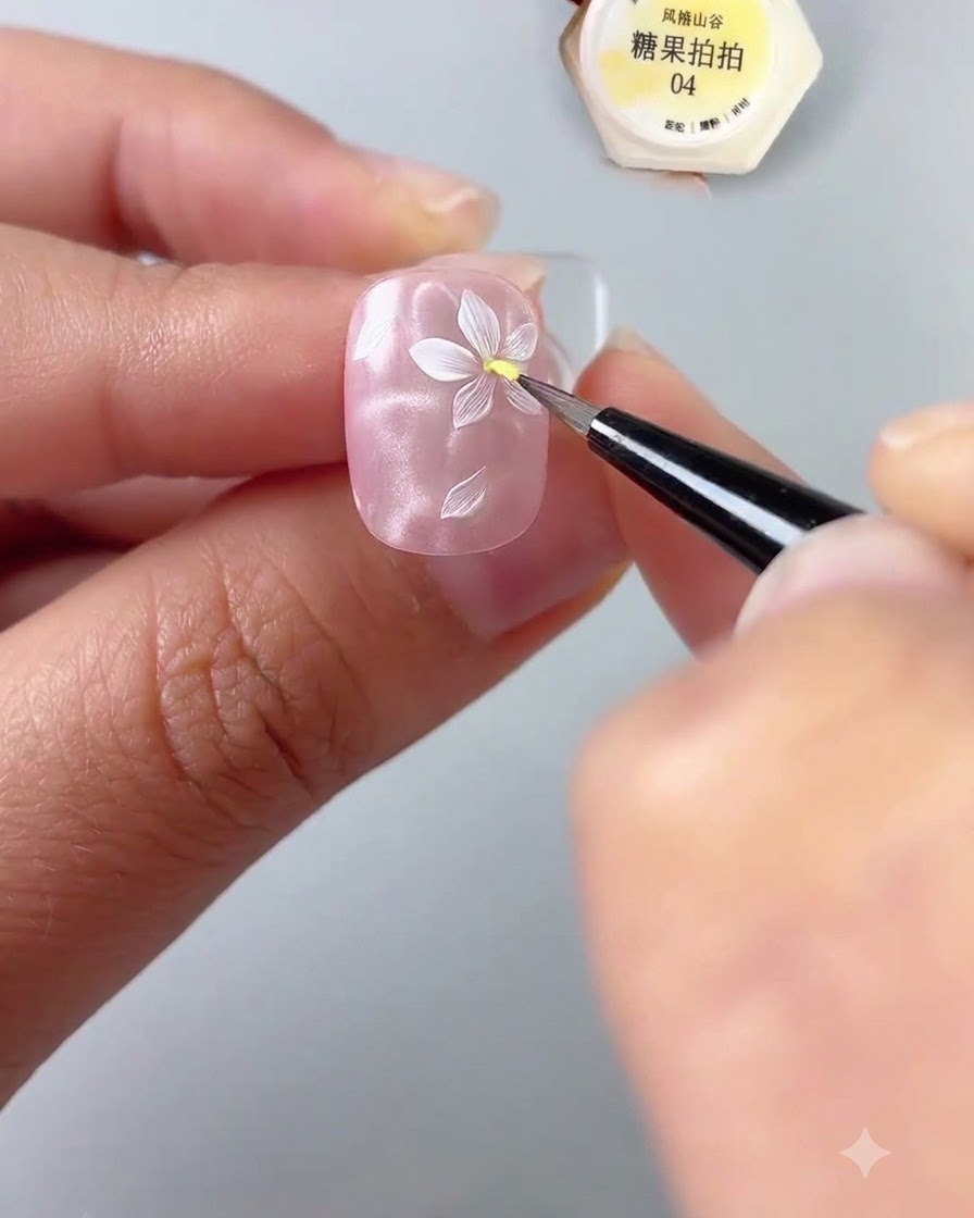 Un primer plano de una mano sosteniendo una uña mientras usa un pincel de detalle fino para aplicar una gota de gel amarillo en el centro de una flor blanca pintada sobre una base de uña rosa brillante.