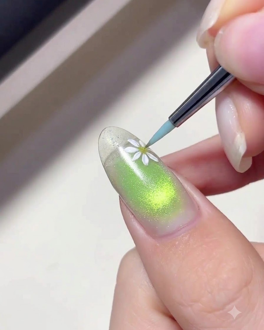 Un pincel delineador fino aplicando gel blanco para pintar delicados pétalos de margarita en una uña con degradado verde.
