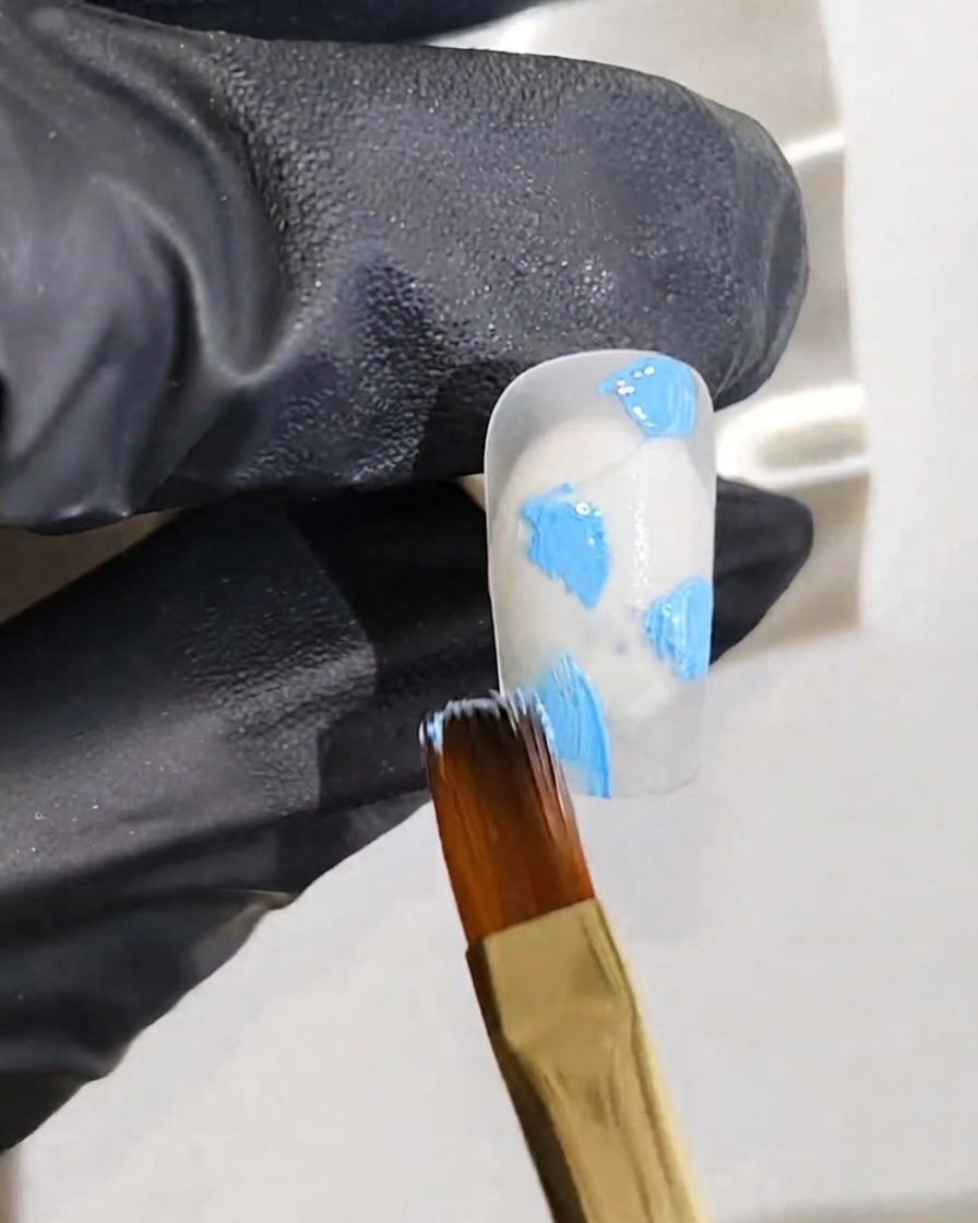 Una mano con guantes sosteniendo un pincel fino aplicando puntos de gel azul sobre una base de uña blanca.